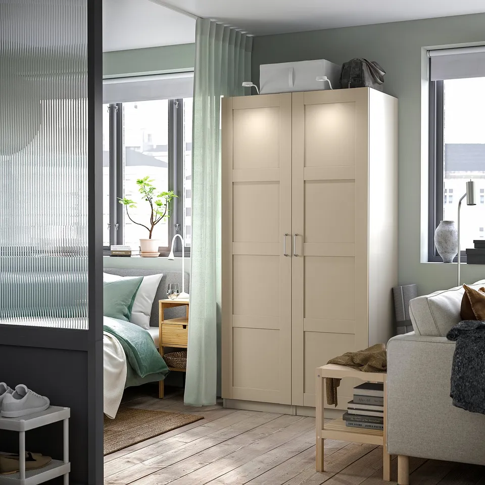 Modern bedroom featuring beige PAX wardrobe with integrated lighting, light wood flooring, blue-green curtains, and cosy decor.