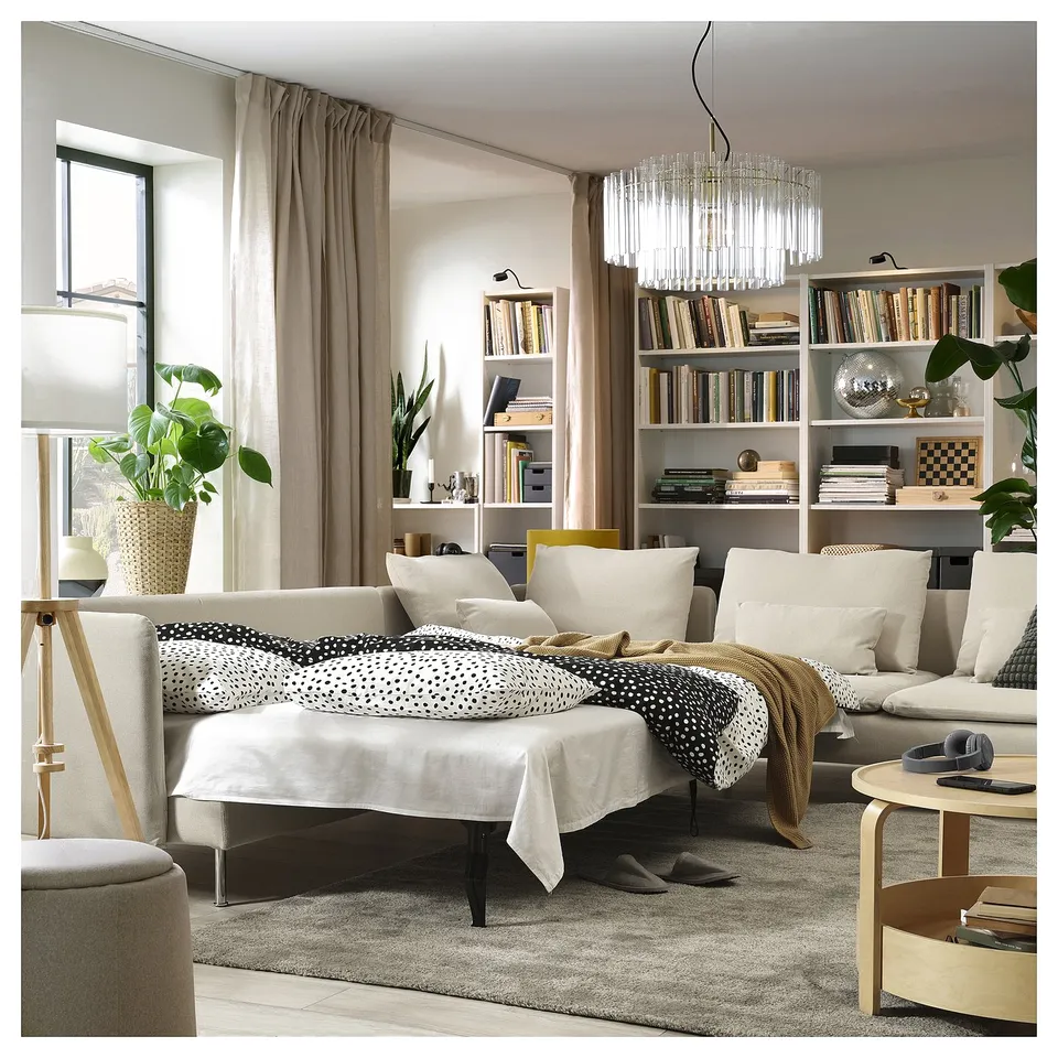 Modern living room with beige SÖDERHAMN sofa bed, white cushions, and polka dot blanket. Bookshelves, plants, and chandelier, cosy and stylish.