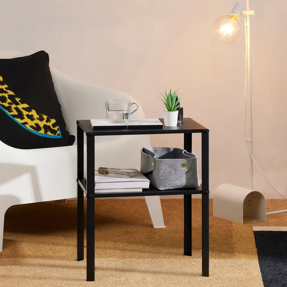 A small black side table with a shelf holding a plant, cups, and books. Brightened by a nearby lamp, ideal for indoor use and easy to move.