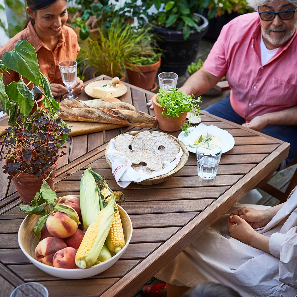 Family enjoys outdoor meal on NORRMANSÖ table. Wooden chairs, fresh food, potted plants.