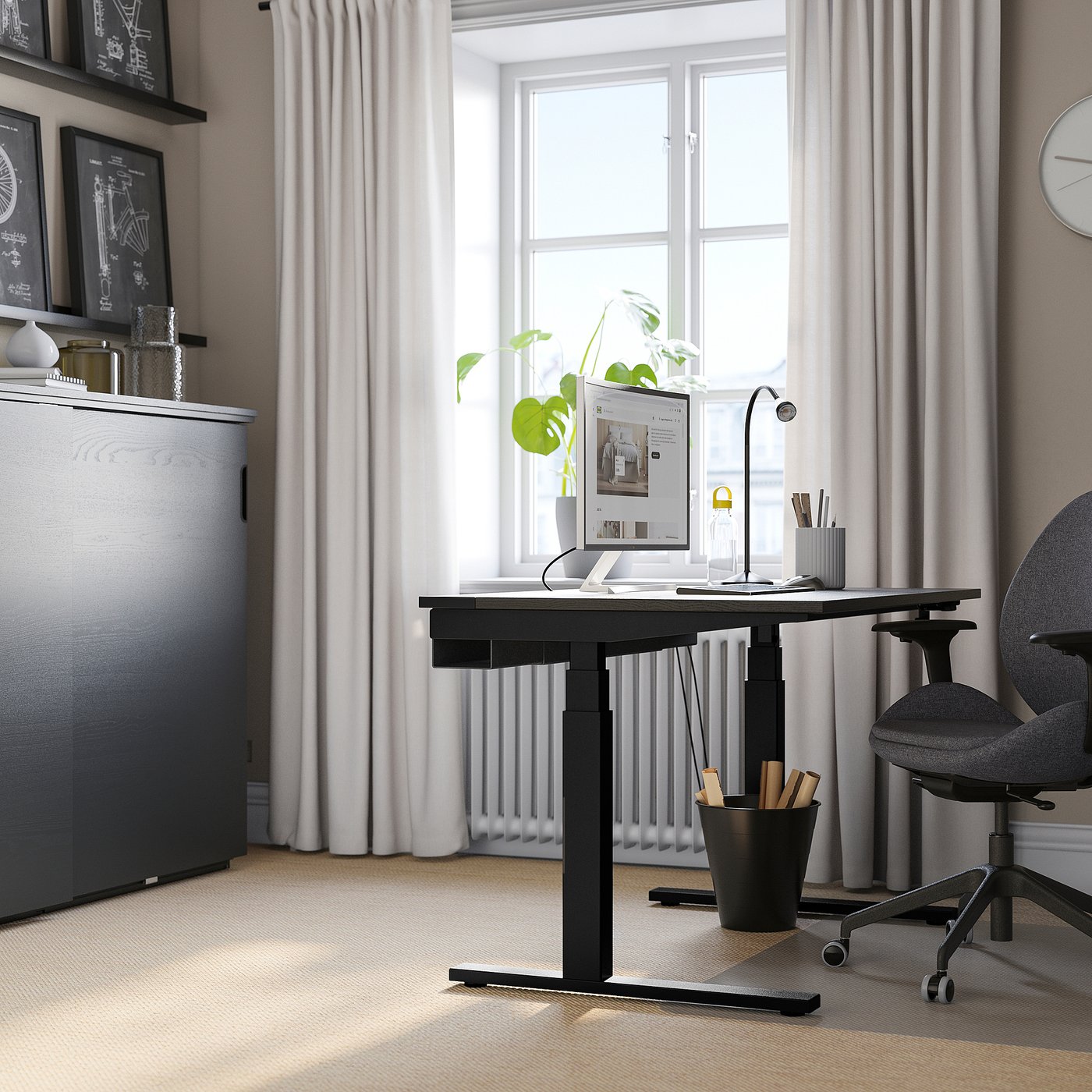 Modern home office with MITTZON black sit-stand desk, grey chair, large windows, white curtains, and grey carpet. Desk holds pc, lamp, and plant.