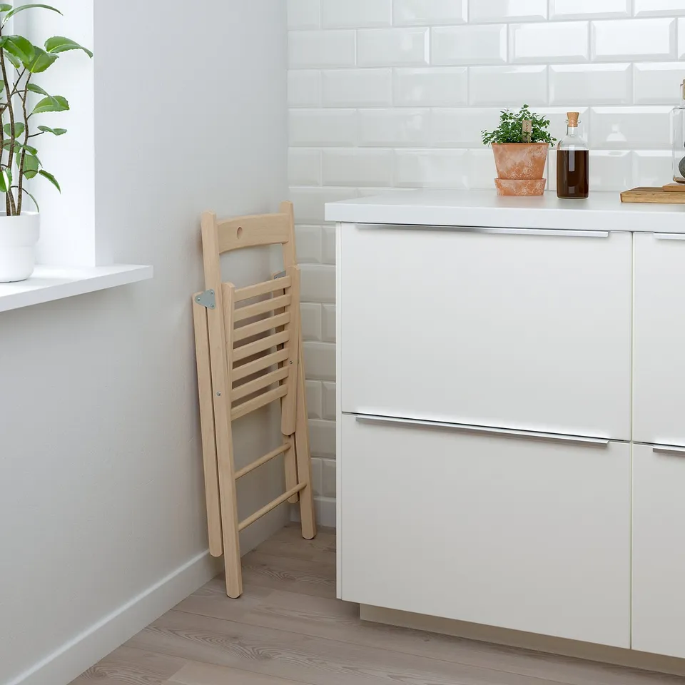 Folding wooden chair leans against white cabinet in minimalist kitchen. Light wood, simple design.