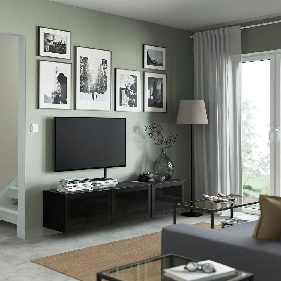 Modern living room with BESTÅ tv bench, flat-screen tv, decorations, grey sofa, glass table, large photos, floor lamp by window.