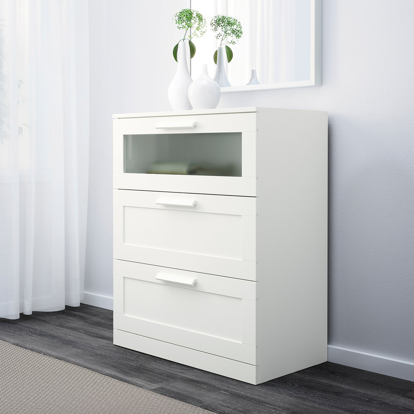 A white chest of drawers with three sections, one with glass. On top are two vases with greenery, set against a white wall and curtain.