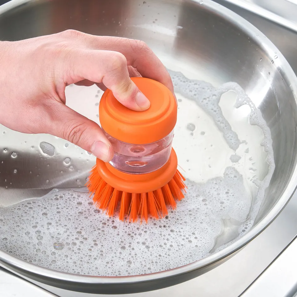 A person holds an orange dish brush over a soapy sink. The brush releases soap from its handle.