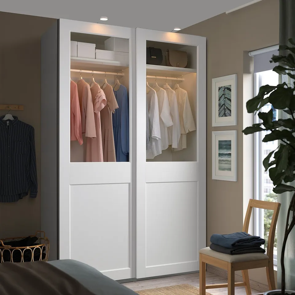 White PAX wardrobe with glass doors, lights, clothes, beside window & chair.
