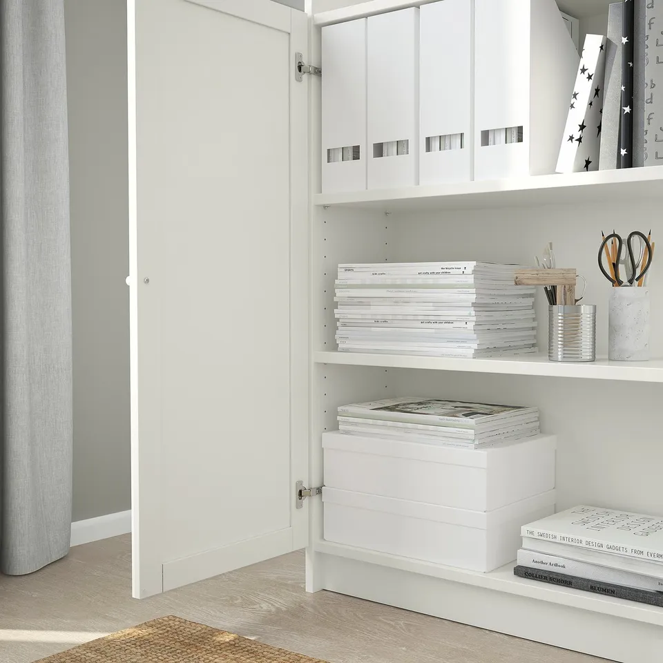 White BILLY bookshelf with adjustable shelves and hinges, displaying books and office supplies, positioned next to a curtained window.