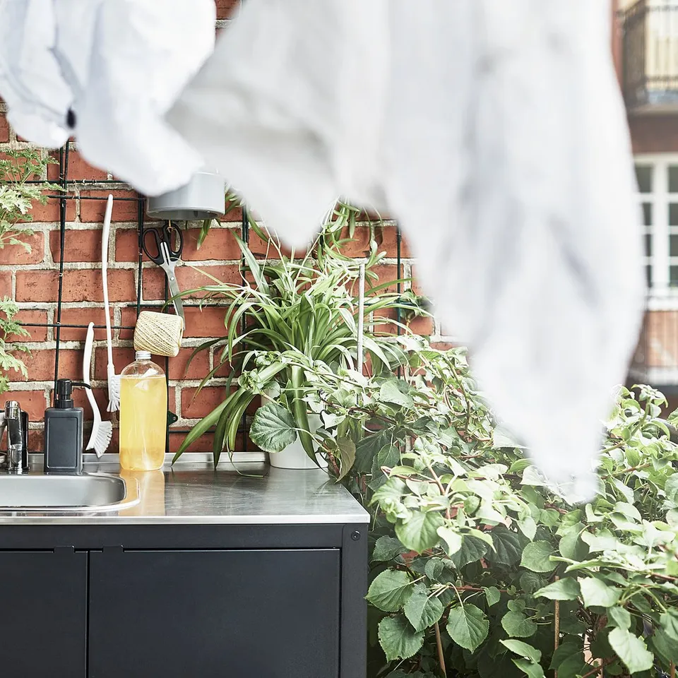 Outdoor sink area with plants. Stainless steel sink on black GRILLSKÄR unit. Hanging towels, green plants, cleaning tools, against brick wall.