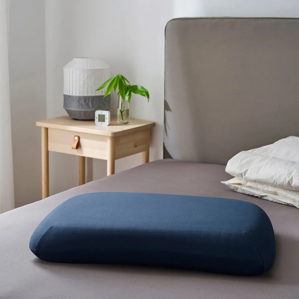 Bed with grey bedding, pillow, light, plant, and clock on bedside table.