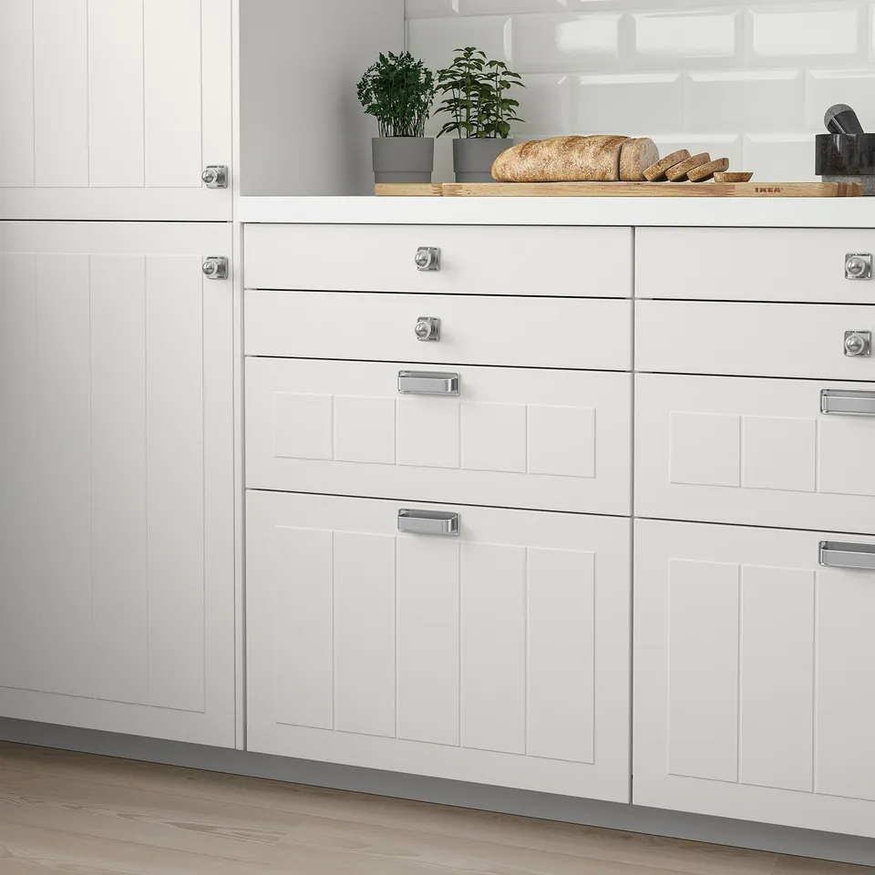 Modern white kitchen cabinets with square metal handles, potted plants, and bread on a wooden board.