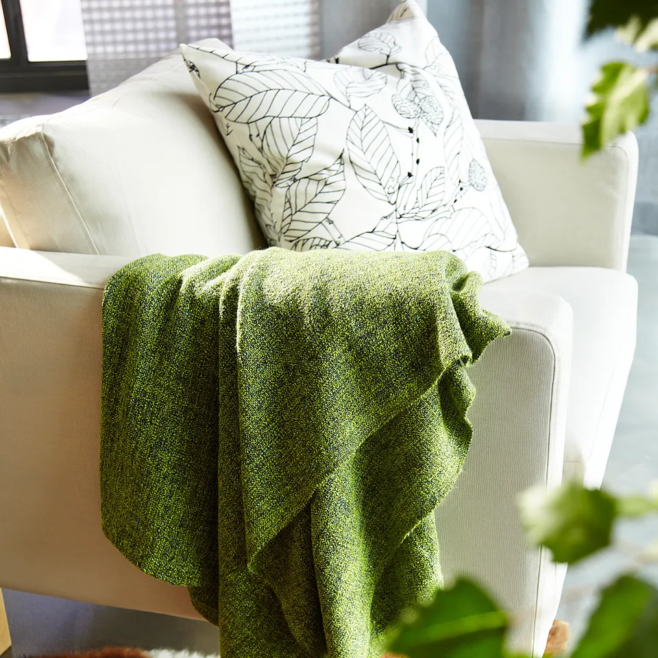 A cosy white chair with a green blanket and a pillow with leaf designs.