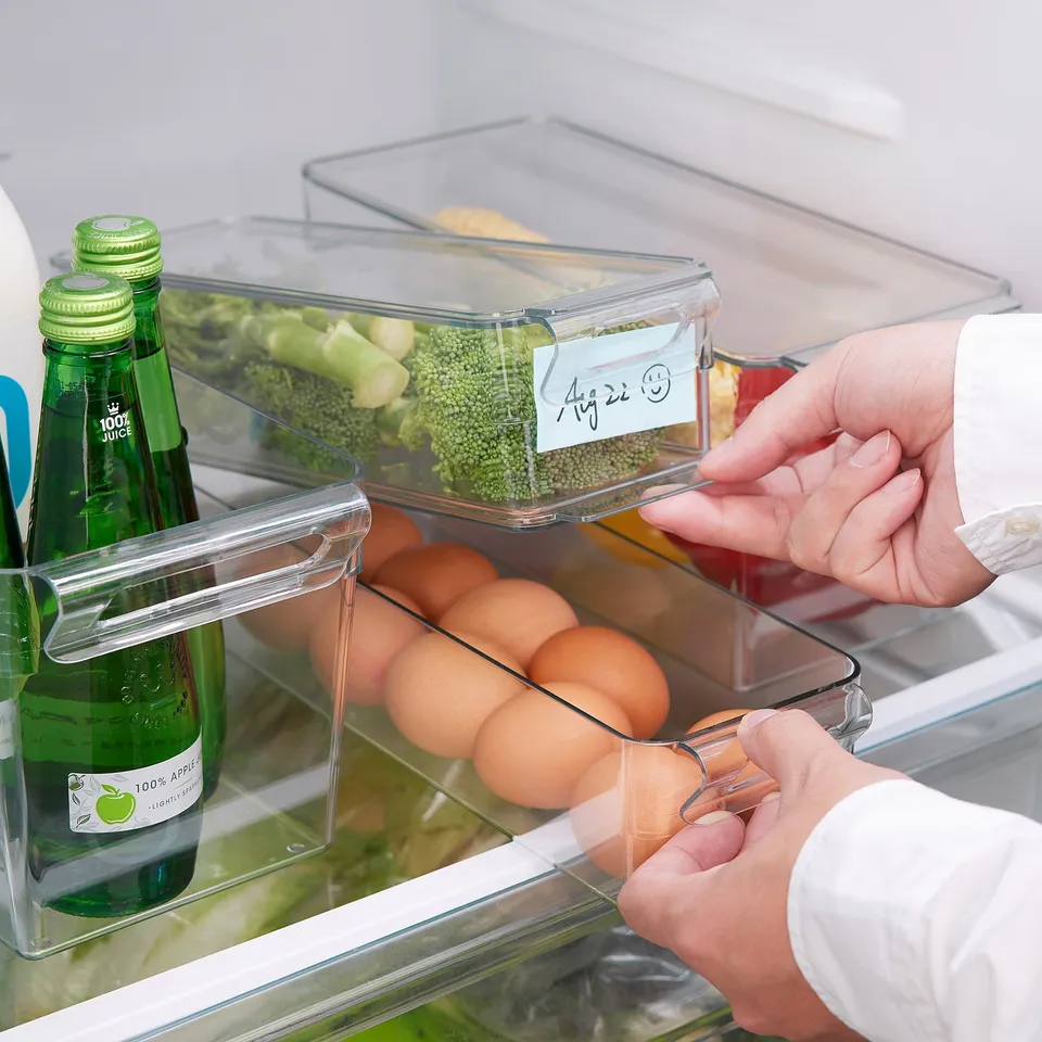 Person organises fridge using clear, stackable KLIPPKAKTUS boxes. Labels indicate stored dates for freshness.