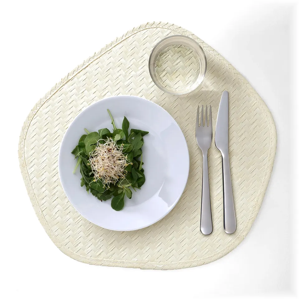 A handwoven palm leaf place mat displaying a salad, knife, fork, and glass, protecting table surfaces and adding natural decor.