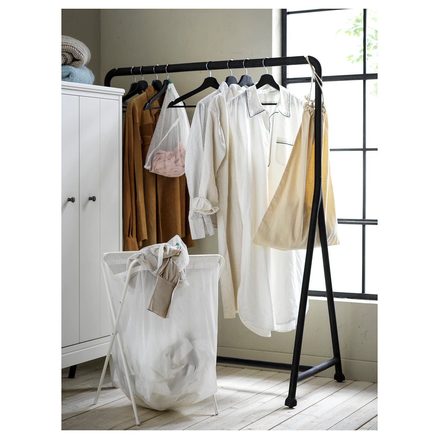 Black metal clothing rack with wheels, holding light-colored clothes near a white cabinet and a white laundry basket.