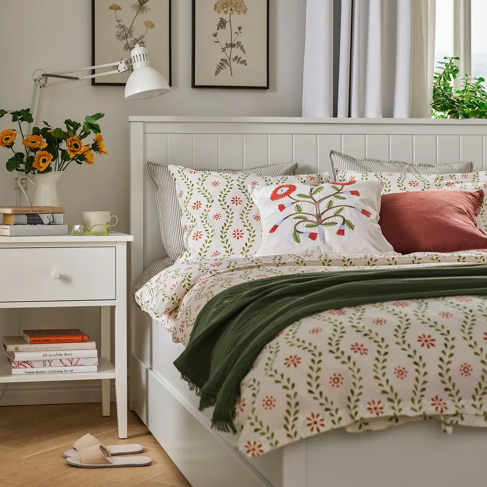White FYRISVIDE bedding with floral patterns on a large bed, beside a table with a lamp, books, and yellow flowers.