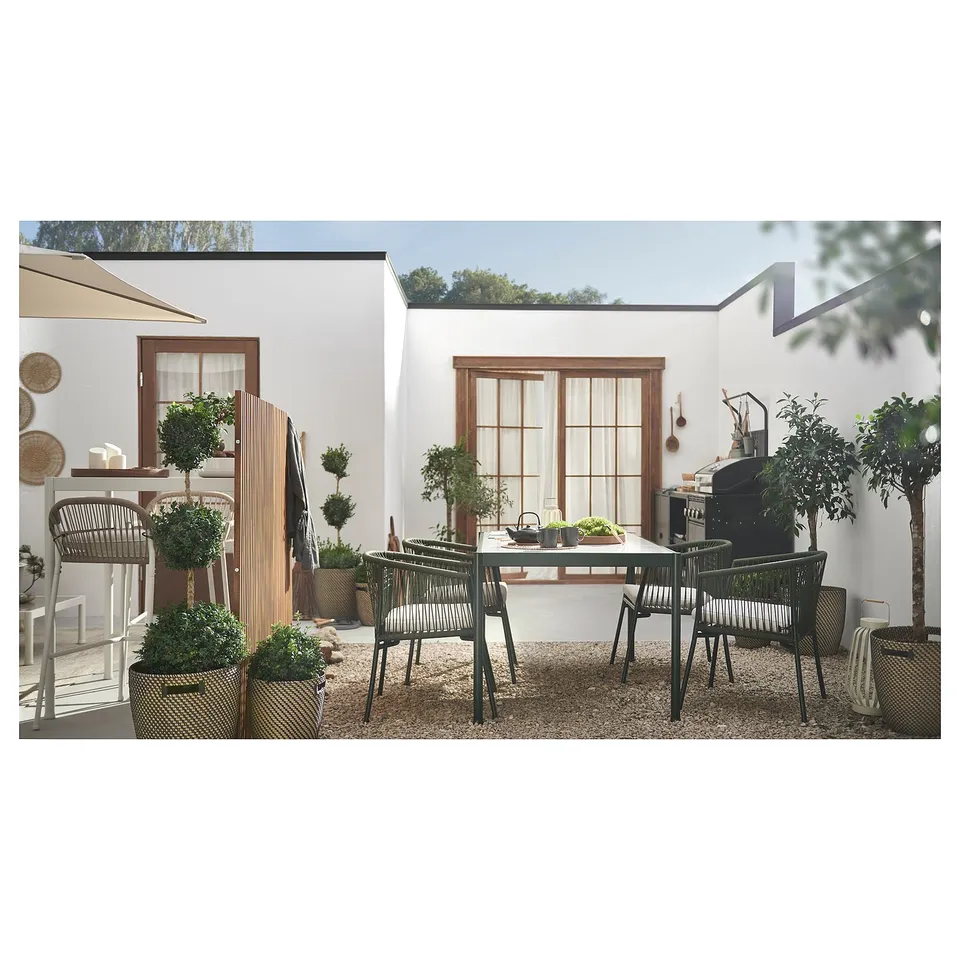 Modern outdoor dining set with wicker chairs and glass table; surrounded by plants, beside a white building.