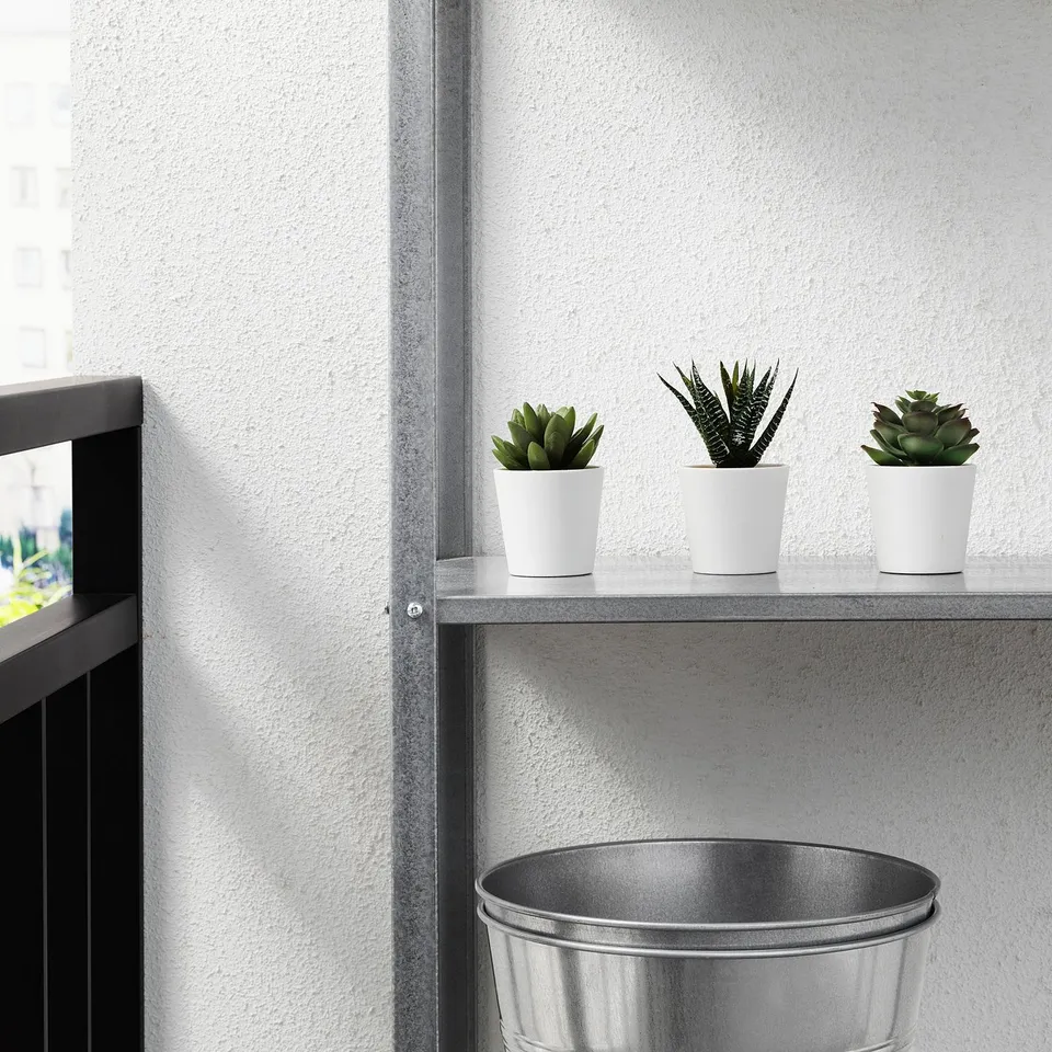 Three artificial succulents in white pots line a modern shelf against a white wall, complementing a sleek metal bucket below.