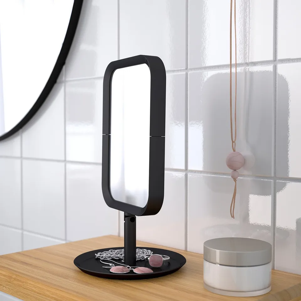 Black vanity mirror with magnification; sits on wooden surface, reflecting light, with jewellery displayed beneath.