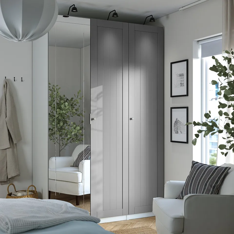 Minimalist bedroom with white PAX wardrobe, mirror door, light wood interiors. White armchair by bed with blue/white bedding, city view window.