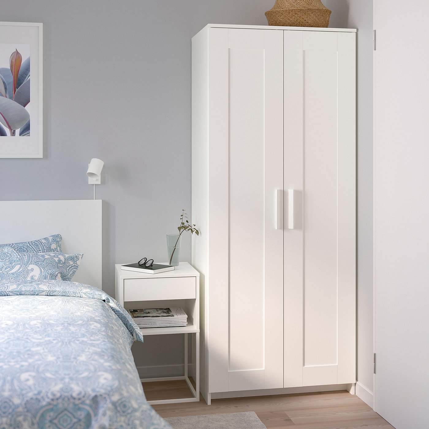 A modern, white BRIMNES wardrobe, tall and narrow, placed beside a bed. Its simple, clean design complements a minimalist bedroom decor.