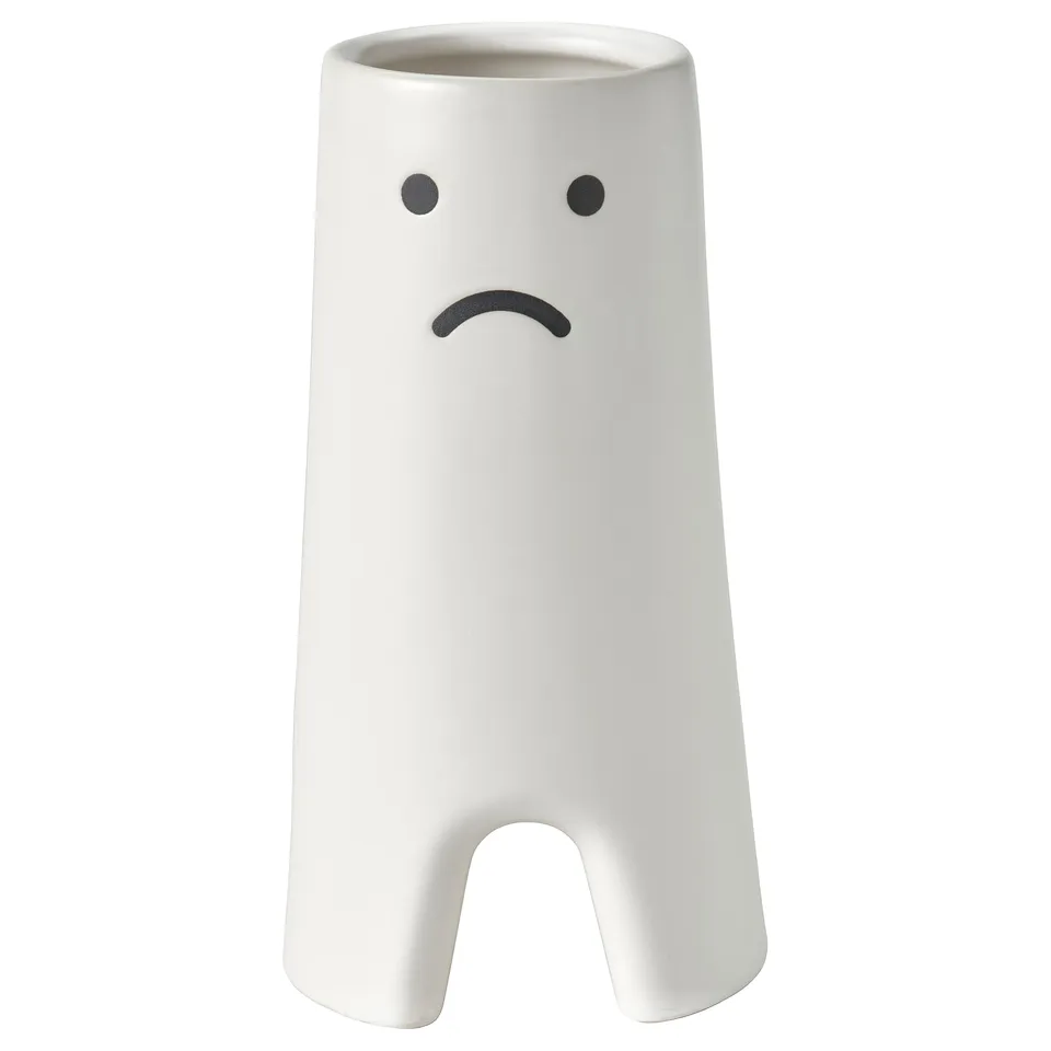 A white, cylindrical vase named FRÖVÄXT with a sad expressive face design. Its made of stoneware and has an opening at the bottom.