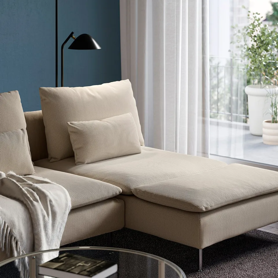 Modern sectional sofa with cushions, a blanket, and a lamp in a minimalist living space. Large windows let in natural light.