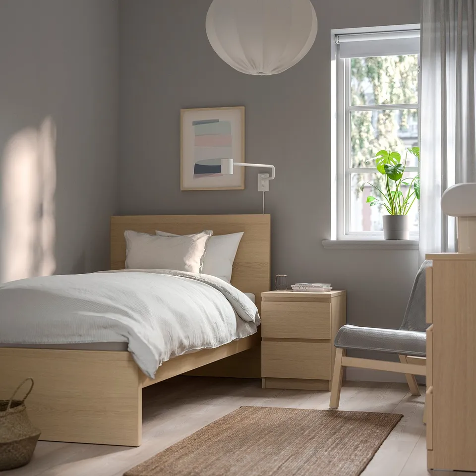 Modern grey bedroom with MALM bed, wooden headboard, white linens, bedside table, chair, window, and plant. Grey walls, light floors, and cosy decor.