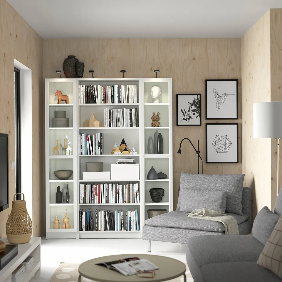Modern living room with white BILLY bookcases, grey sofa, and decorative art frames. Wooden walls, glass doors, and various ornaments.