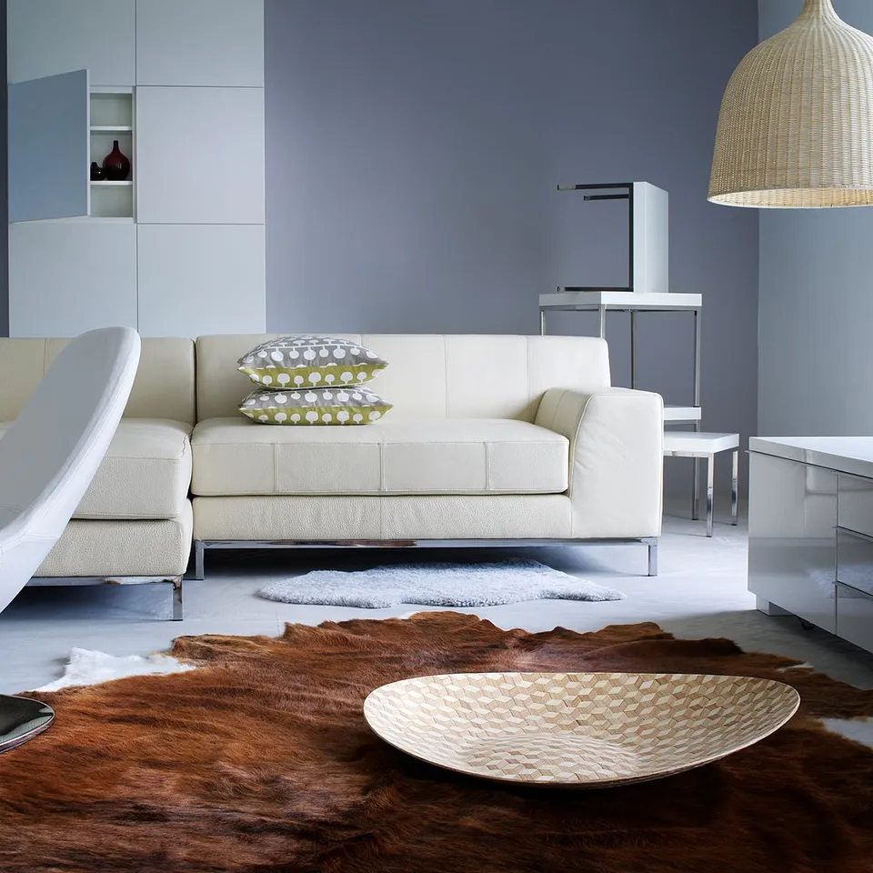 Contemporary living room with white sectional sofa, cowhide rug, white armchair, and woven pendant lamp on blue wall.