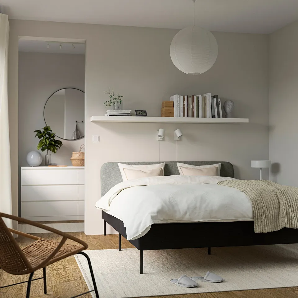 Minimalist bedroom with grey SLATTUM bed, shelves, wicker chair, and pendant light. Soft rug and slippers add comfort.