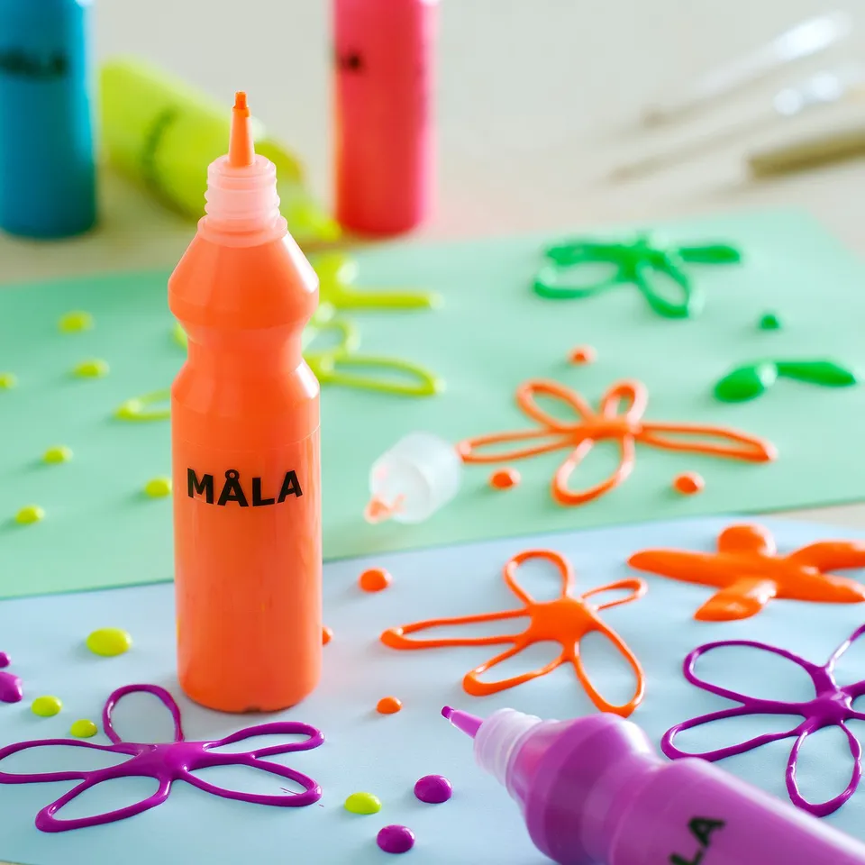 Bottles of non-toxic MÅLA kids paint in bright colours displayed on a green surface, some with painted flower designs.