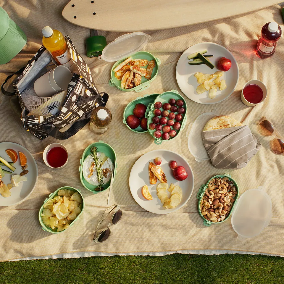 A picnic spread on a blanket with sandwiches, fruits, snacks, and utensils, including green stackable lunch boxes and a hat.