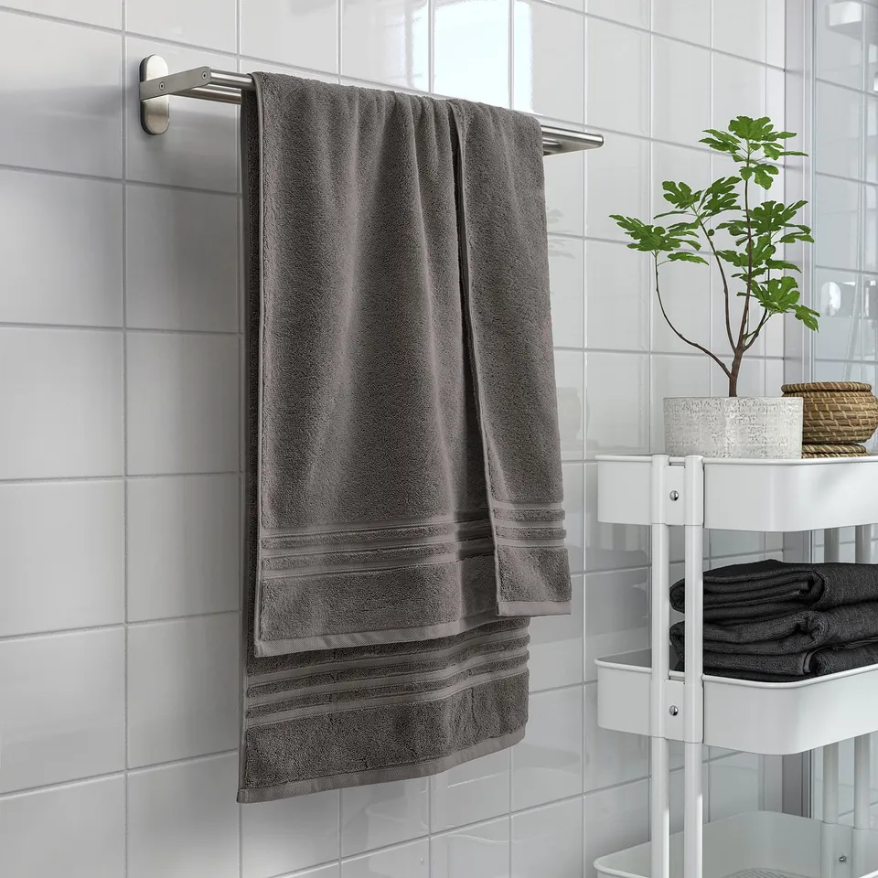 Grey organic cotton BROKGLIM towel with woven border, metallic hanger, and nearby shelf with towels and plant.