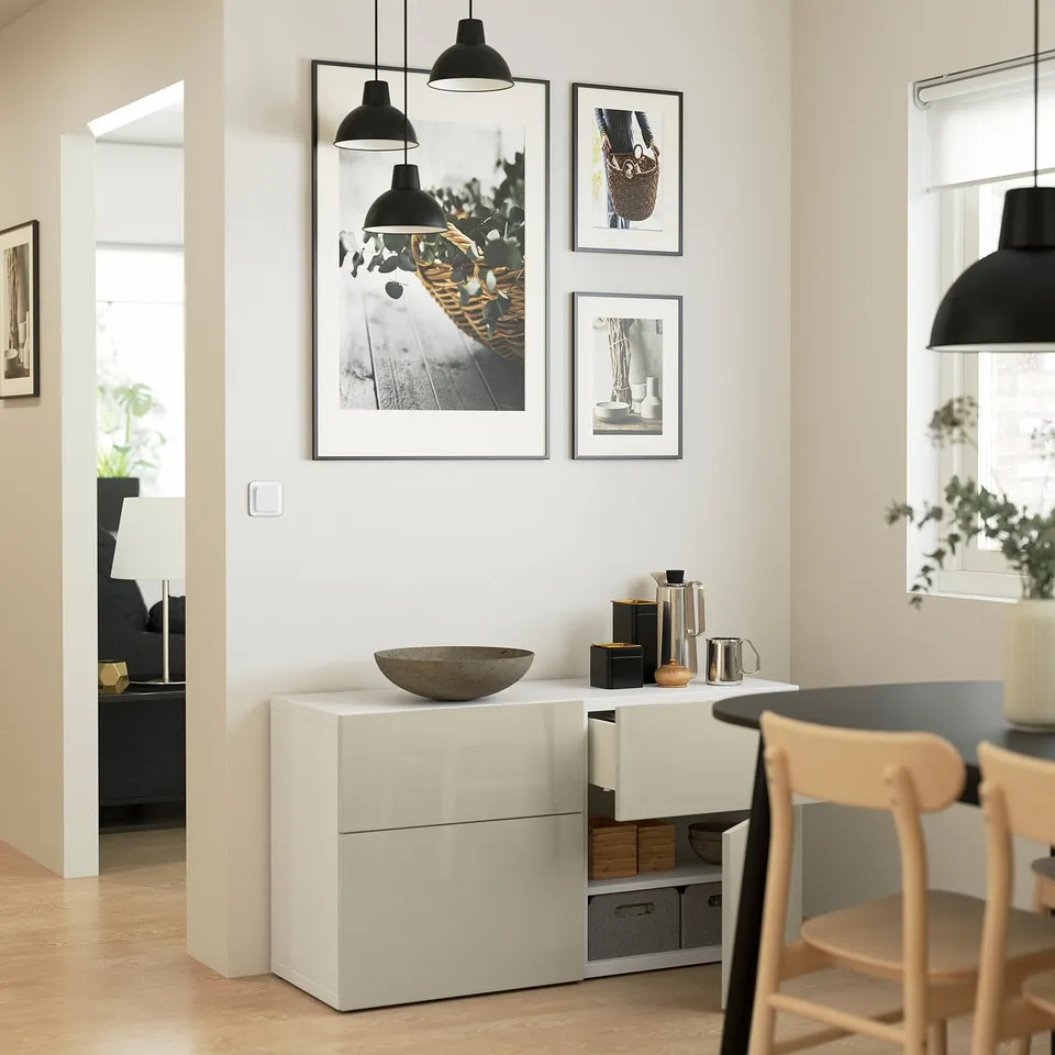 Modern dining room featuring white storage unit with glossy surface, hanging decorative lights, framed art, and wooden chairs.