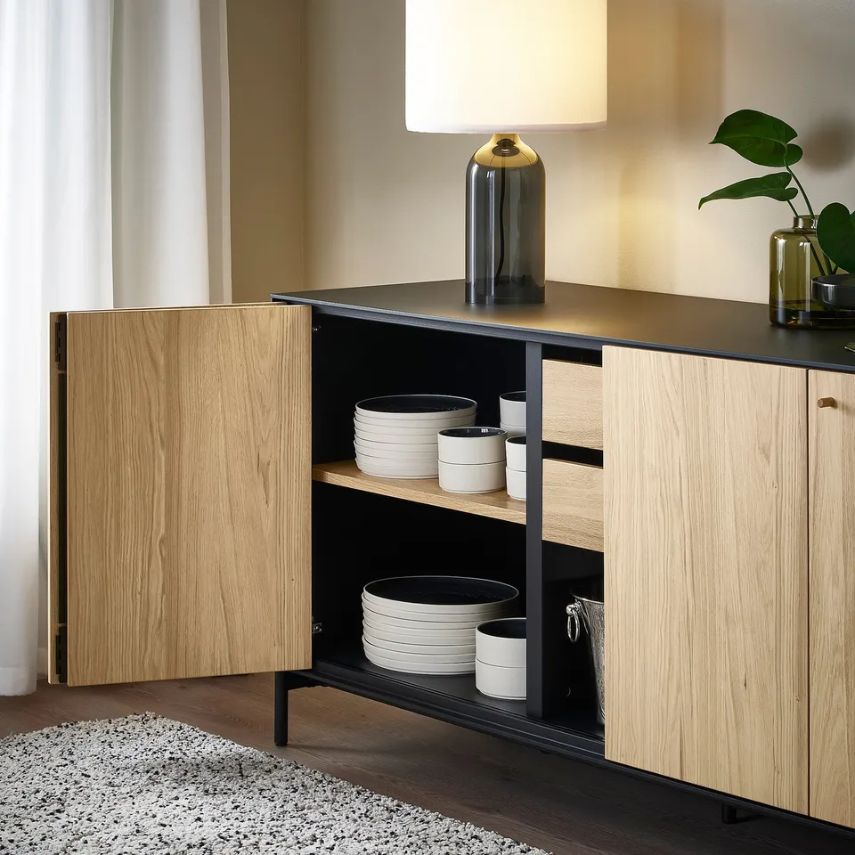 Wooden sideboard with black accents, one door open showing stacked white dishes and cups; stylish, modern design.
