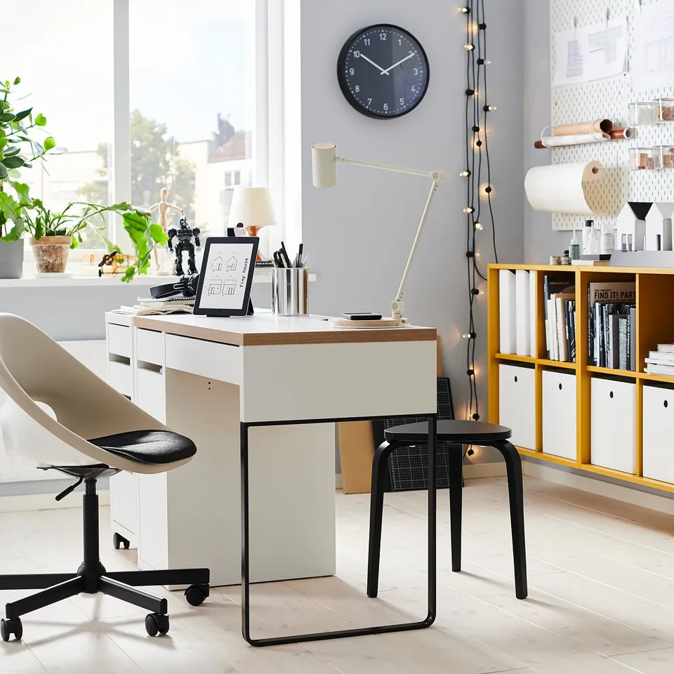 Modern home office with ELDBERGET black chair, white desk, wooden stool, plant, clock, and bookshelf with books and decorations.