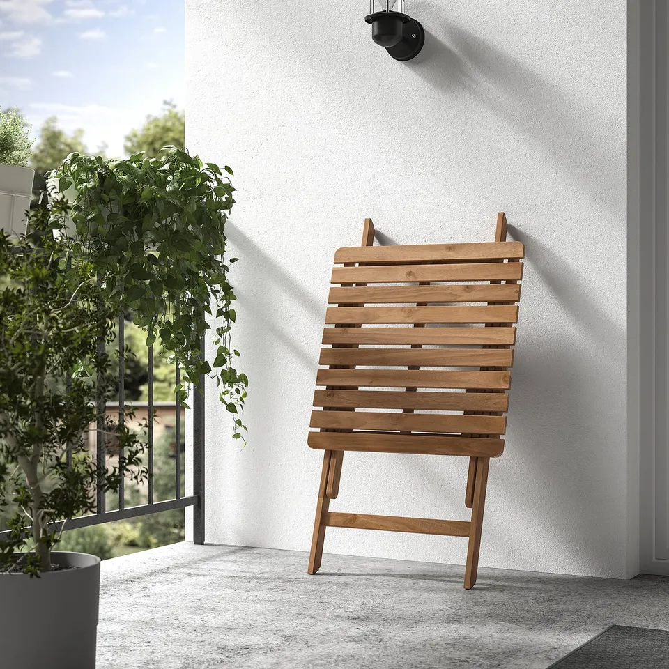 Wooden chair against white wall. Positioned on balcony, with hanging plant nearby. Light-colored wood and simple design.