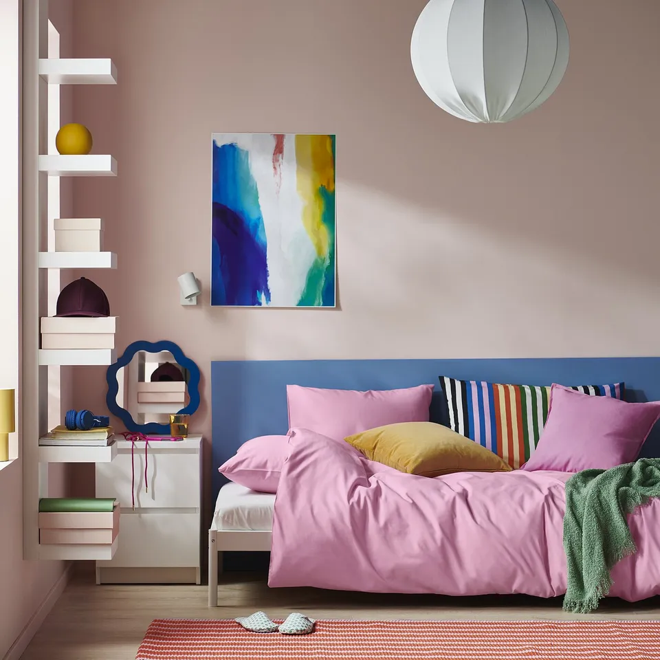 Bedroom with bright pink PILTANDVINGE duvet cover, striped pillows, floating shelves, & abstract wall art. Light grey pendant.