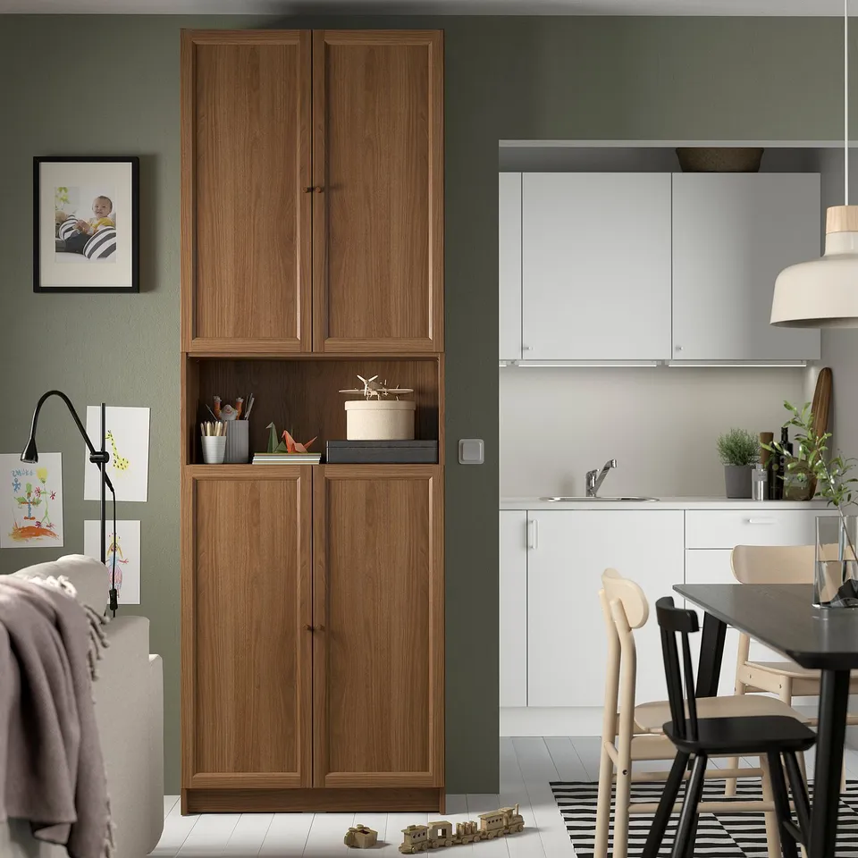 BILLY walnut bookcase between living area & kitchen. Displays toys & decor. Kitchen has white cabinets.