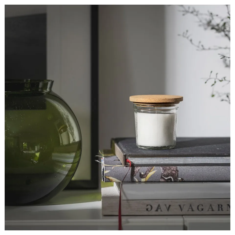 Small round glass jar with white candle and cork lid sits atop books next to green vase.