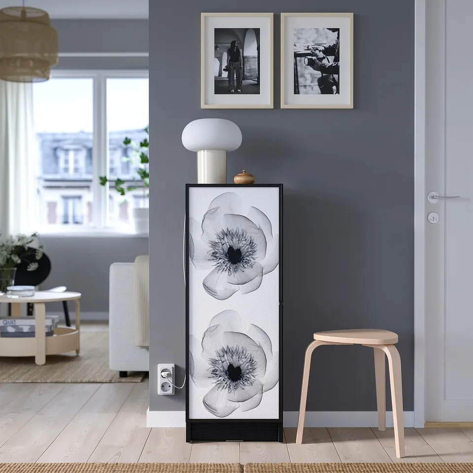 Modern glass cabinet with black-and-white flowers, lamp, and bowl against grey wall.