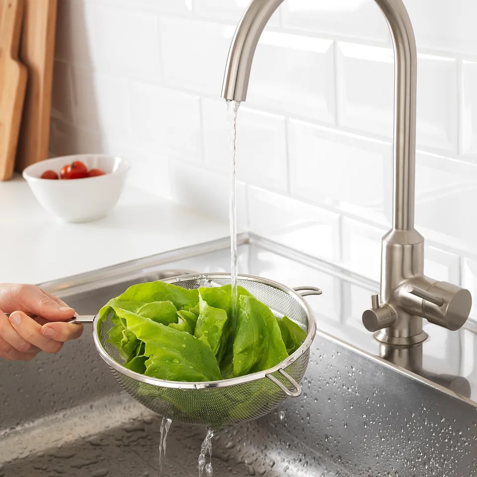 Stainless steel sink with a modern tap. Lettuce is being rinsed in a strainer under running water, indicating its function in washing vegetables.