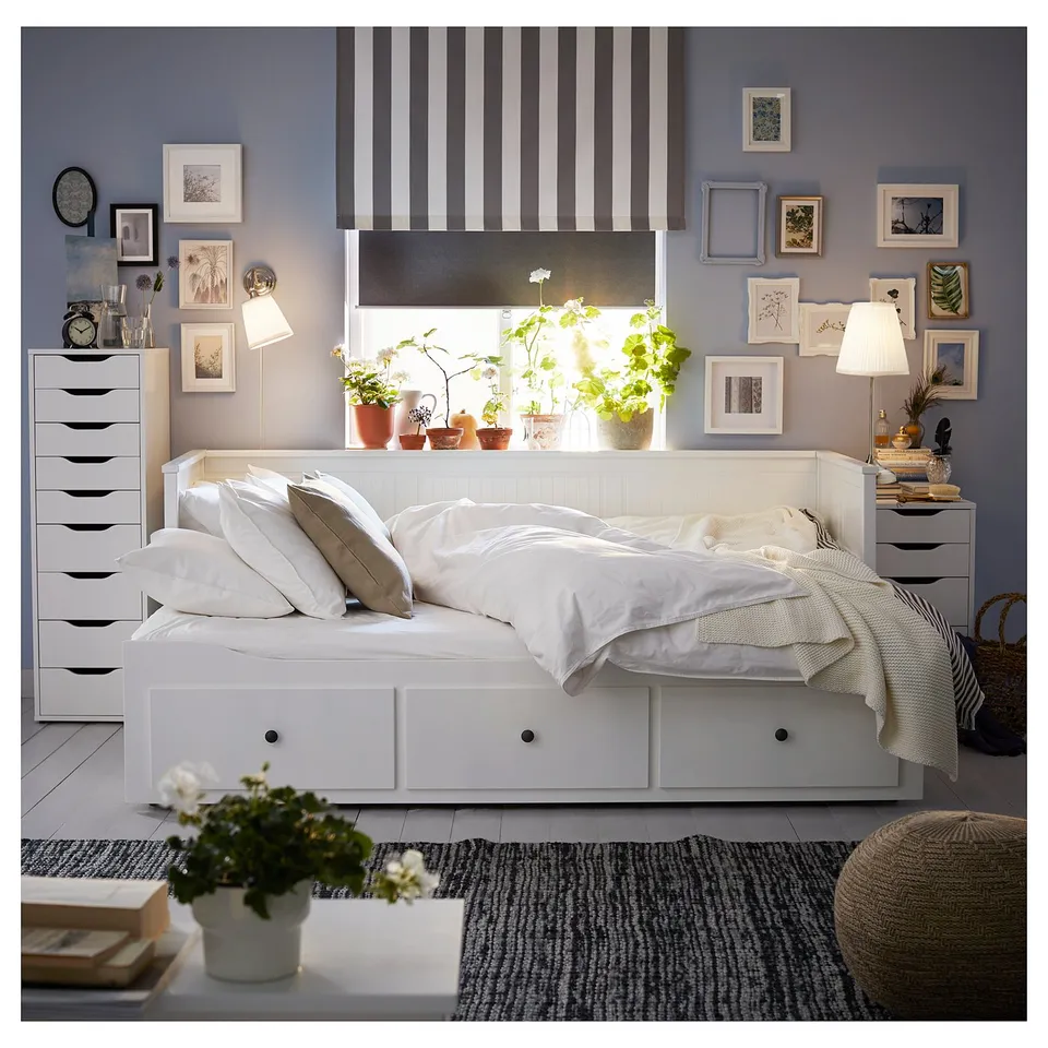 Cosy bedroom with white HEMNES day-bed, striped rug, and blue walls. Greenery by window, pictures on wall, soft lighting.