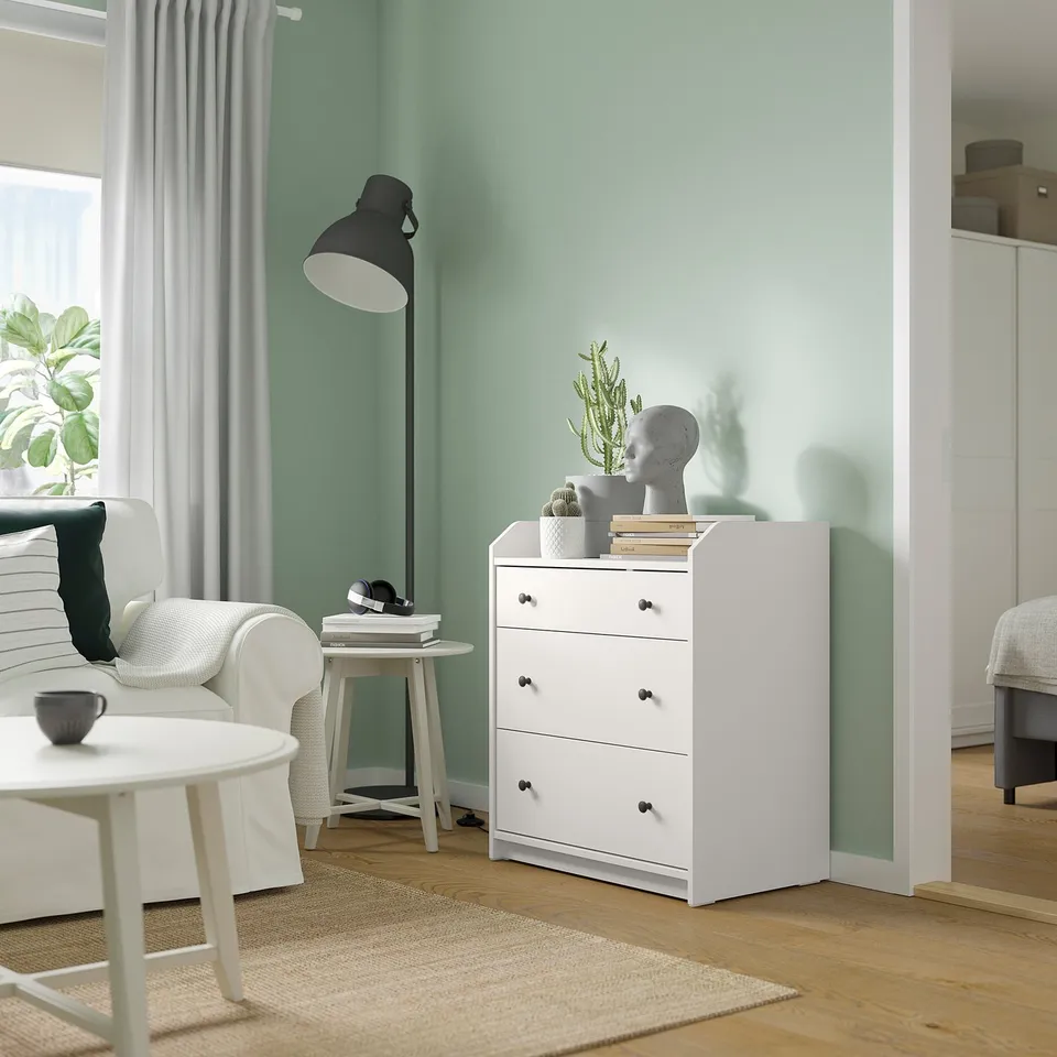 Modern living space with white HAUGA chest, black lamp, light green walls, plush furniture, and a jute rug.