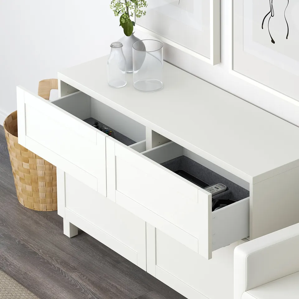 White BESTÅ sideboard with two open drawers revealing remotes, atop wooden floor near wicker basket and white wall decor.