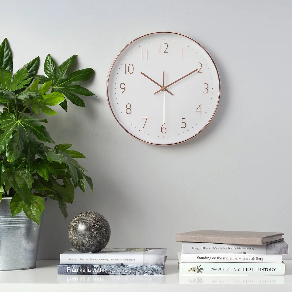 A white wall clock with copper accents displays time on a grey wall, flanked by a potted plant and stacked books on a shelf.