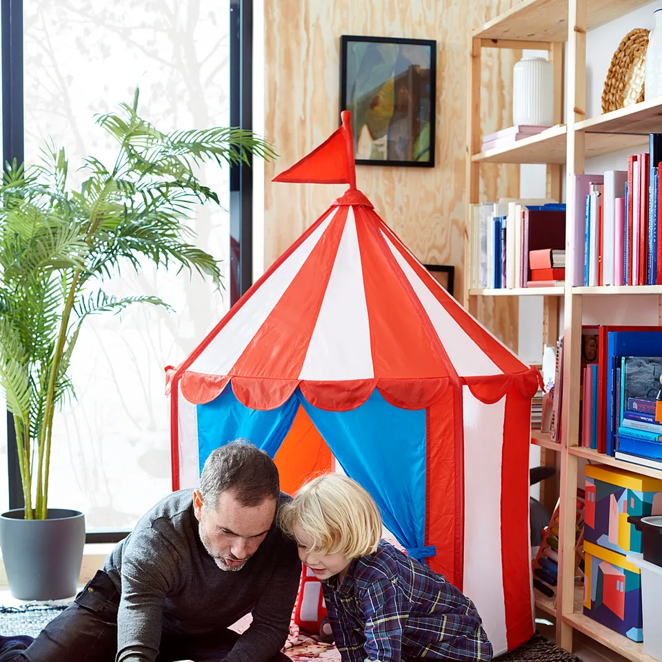 Adult and child playing with wooden train set on rug, with bookshelf and plant.