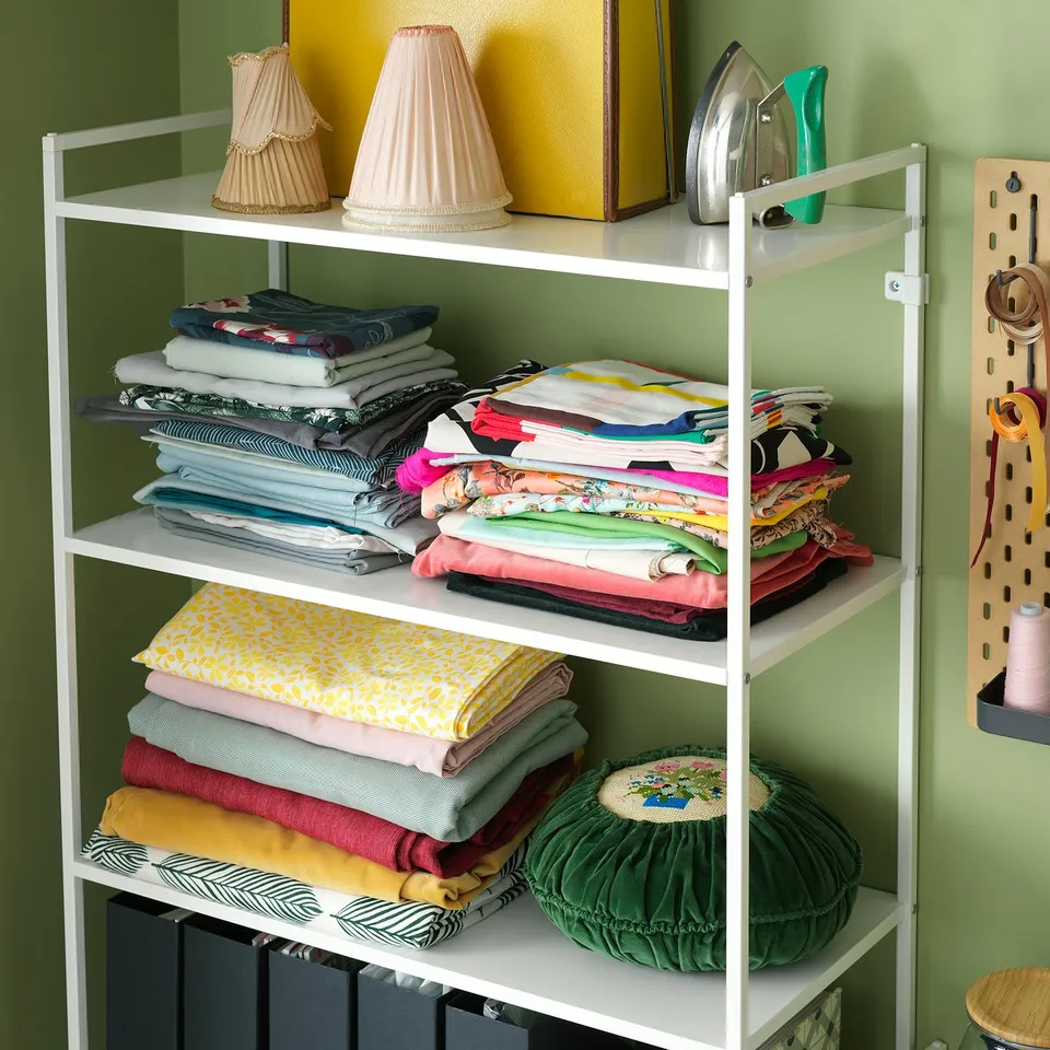 White JONAXEL shelving unit with three shelves holding colourful folded fabrics and decorative items.