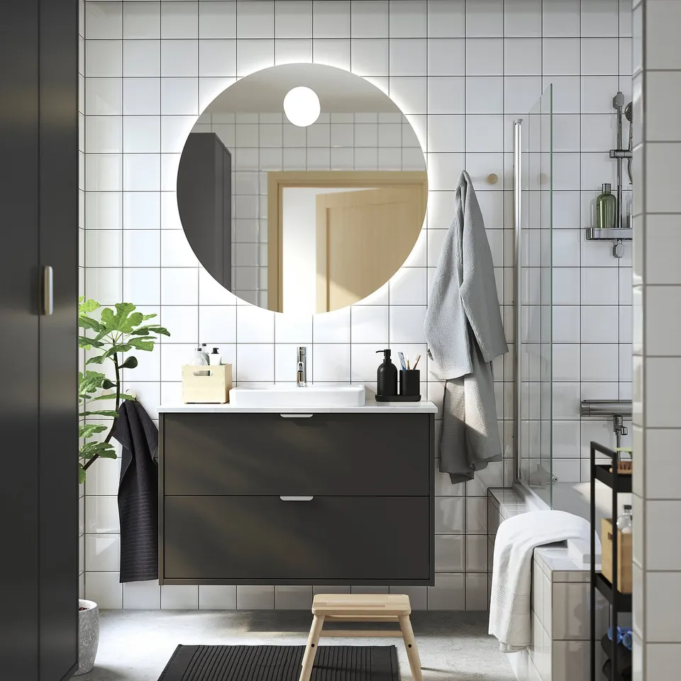 Modern bathroom with dark grey vanity, round mirror, minimalist decor. White countertop, plant, towels, white tiles.