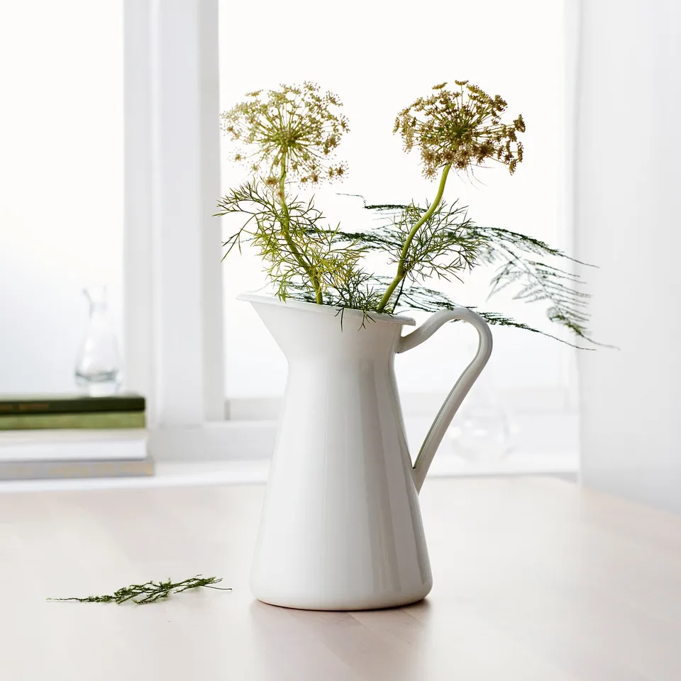White jug-shaped vase with long green stems and lacy flowers. Sits on a light surface near a window.